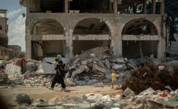 A mother carries a young child in her arms while another young child walks behind her, passing in front of a demolished building in Gaza.