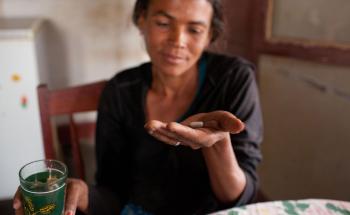 A woman sits at a table, In her hand is medication for HIV/Aids.