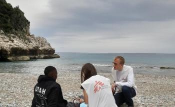 A man is sitting on a beach, catching his breath, while an MSF doctor is examining him. 